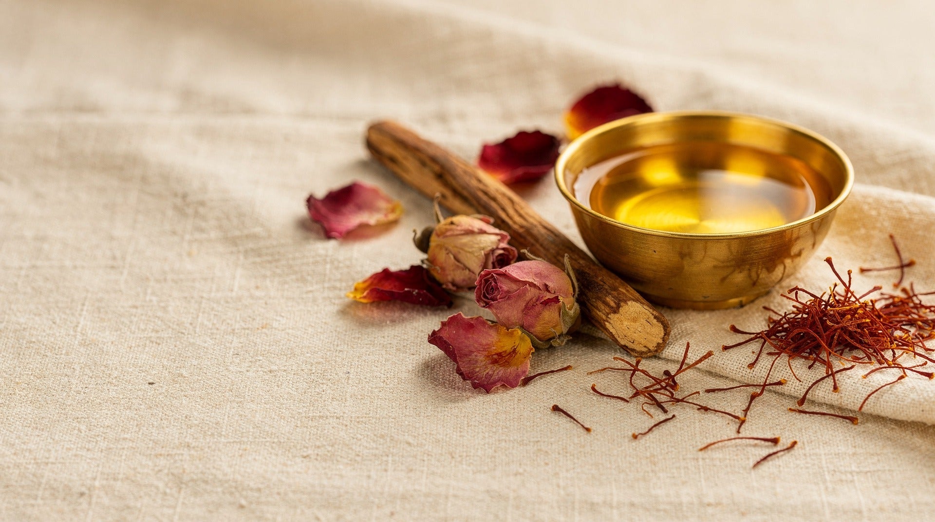 Brass bowl of face oil with rose buds, saffron strands and a sandalwood stick on linen